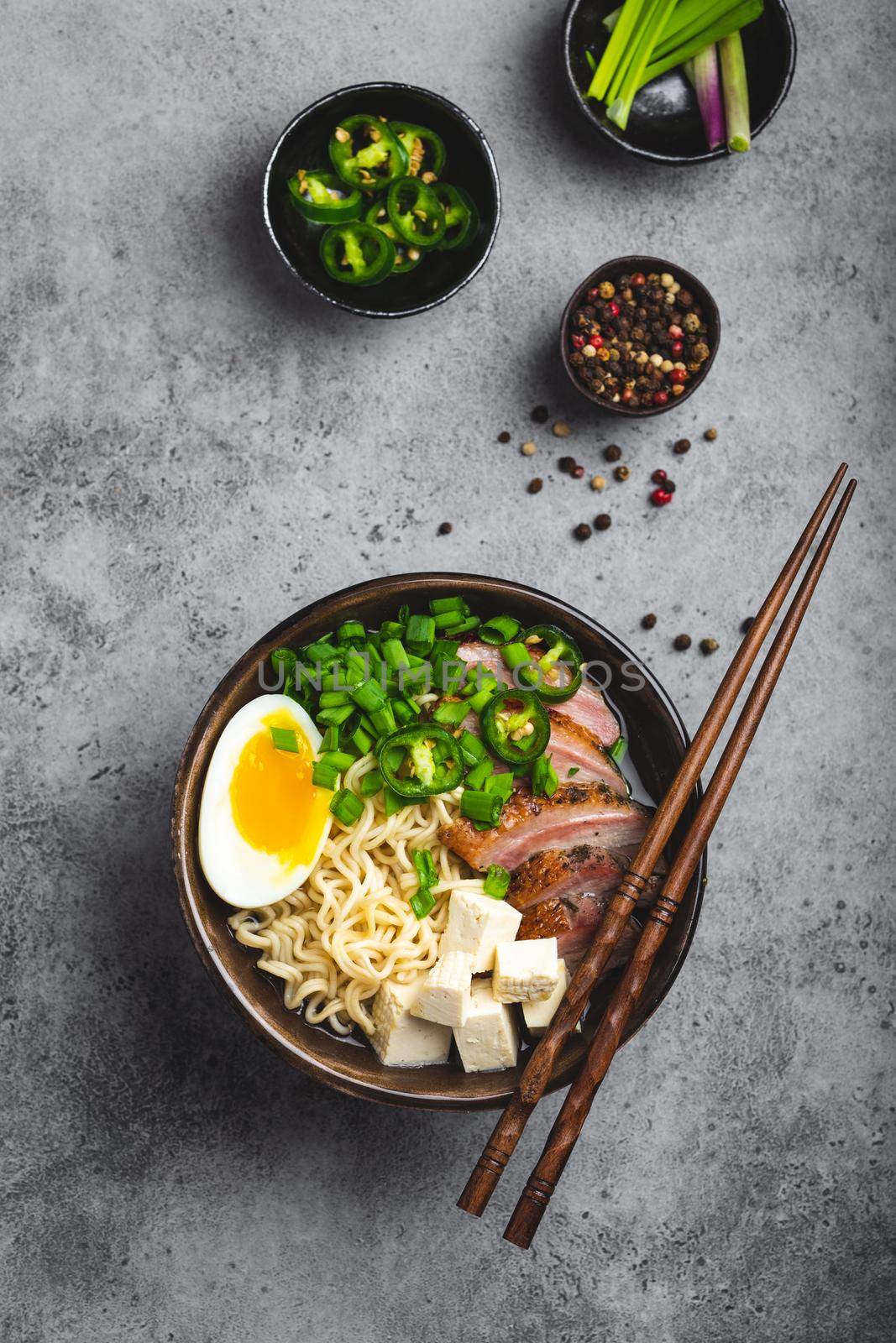 Delicious Asian noodle soup ramen in bowl with meat broth, tofu, sliced pork, egg with yolk, grey rustic concrete background, close up, top view. Hot tasty Japanese ramen soup for dinner asian style