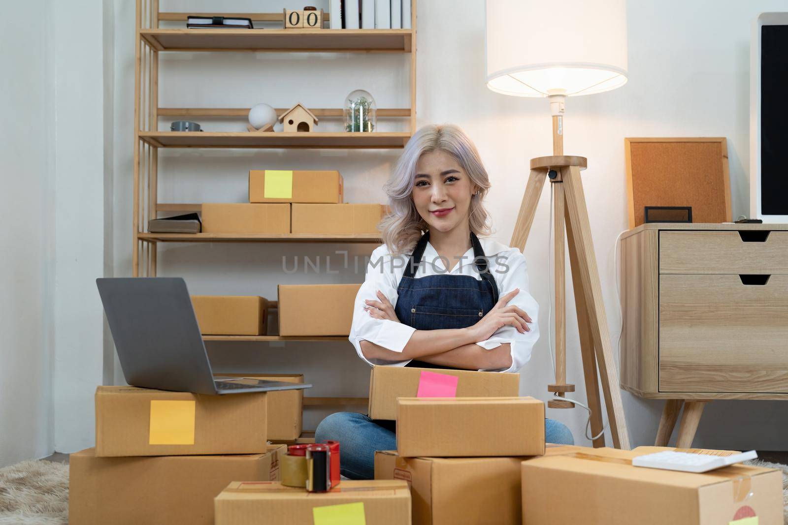 Success young asian woman sit among many boxes or carton also look at layer of boxes with happiness in concept of success sme or e-commerce for new normal lifestyle. by nateemee