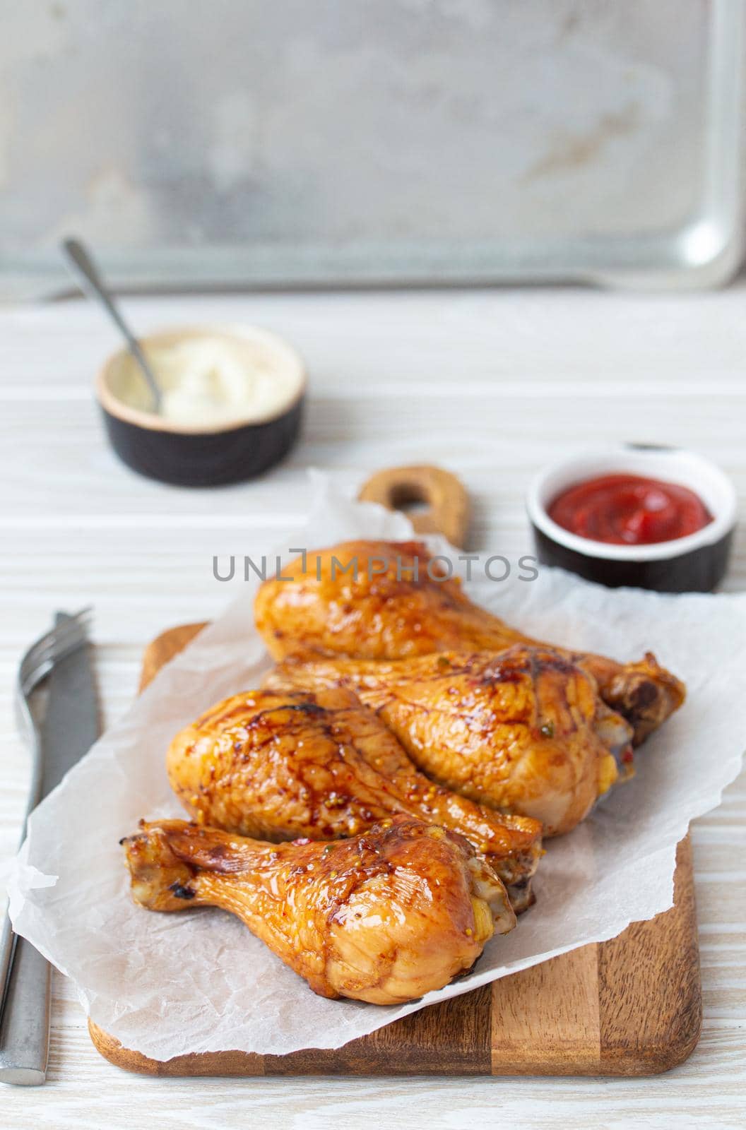 Roasted golden chicken legs drumsticks on baking paper on wooden board served with ketchup and mayonnaise on rustic white wooden table, angle view