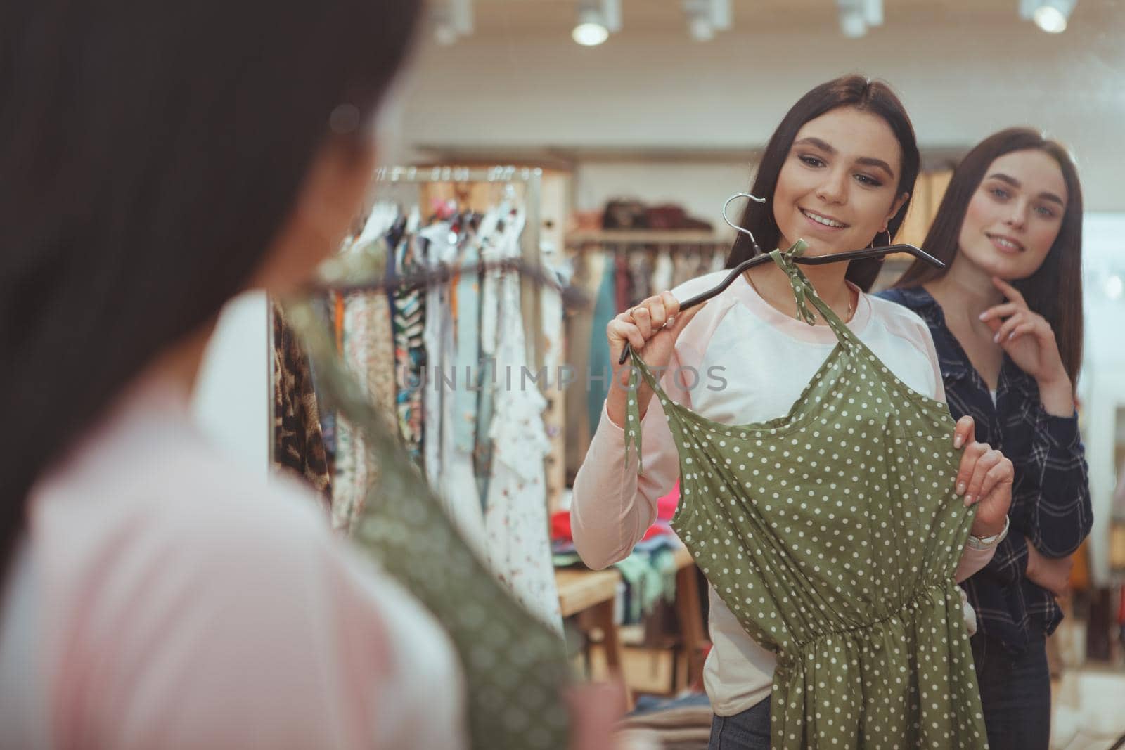 Charming female friends having fun choosing new dress to buy at clothing store. Beautiful woman trying on new dress while shopping with her friend. Friendship, lifestyle, teenagers concept