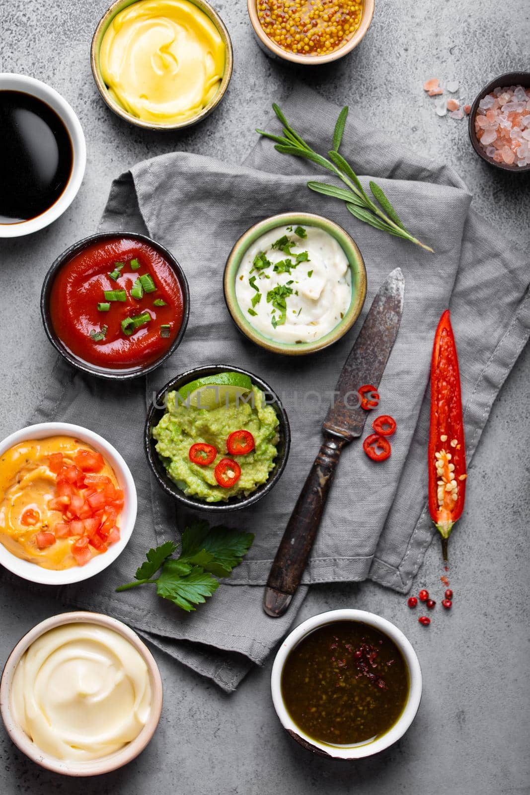 Cooking healthy sauces and dressings concept. Different dips in bowls on rustic concrete background, ingredients, herbs, pepper chili and kitchen knife, top view, close-up