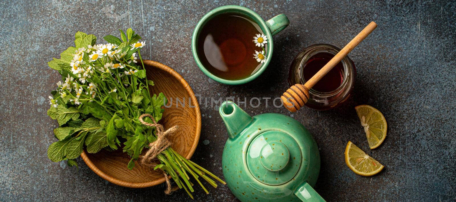 Herbal tea with camomile, fresh medical herbs and flowers, tea cup, tea pot, jar of honey, lemon top view on stone background, herbal alternative medicine and homeopathy remedy concept