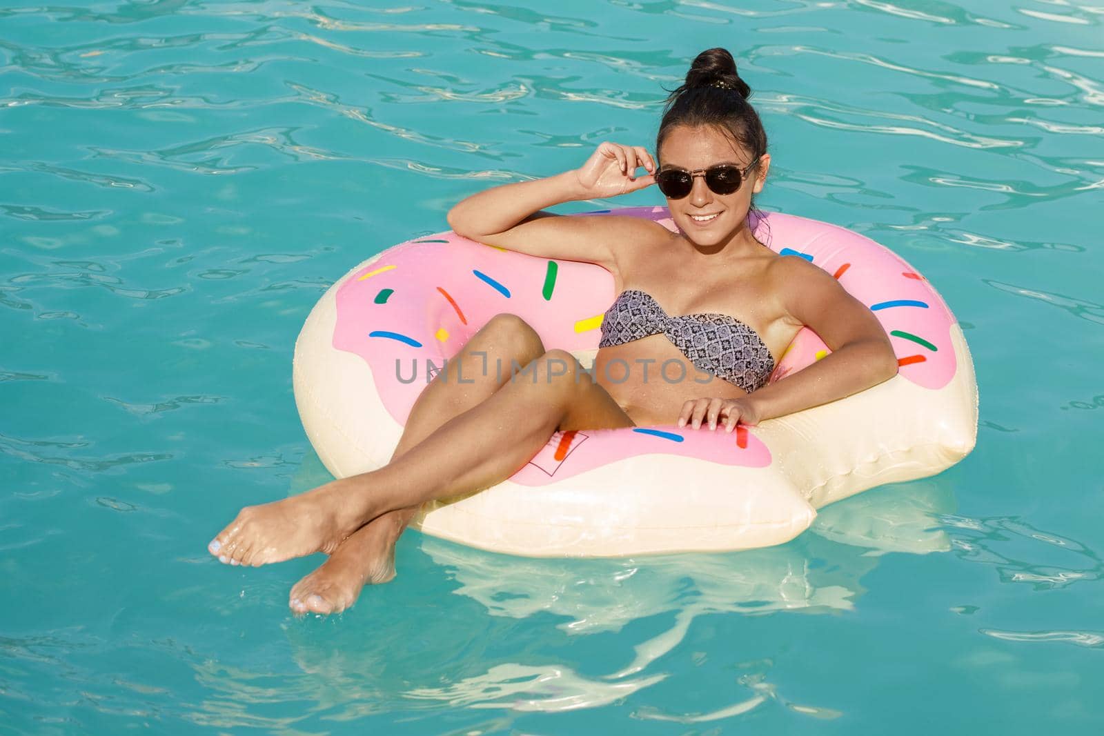 Gorgeous young fit woman smiling to the camera while relaxing at the swimming pool, floating on inflatable donut, copy space. Happy woman enjoying her summer vacation, sun tanning at the pool