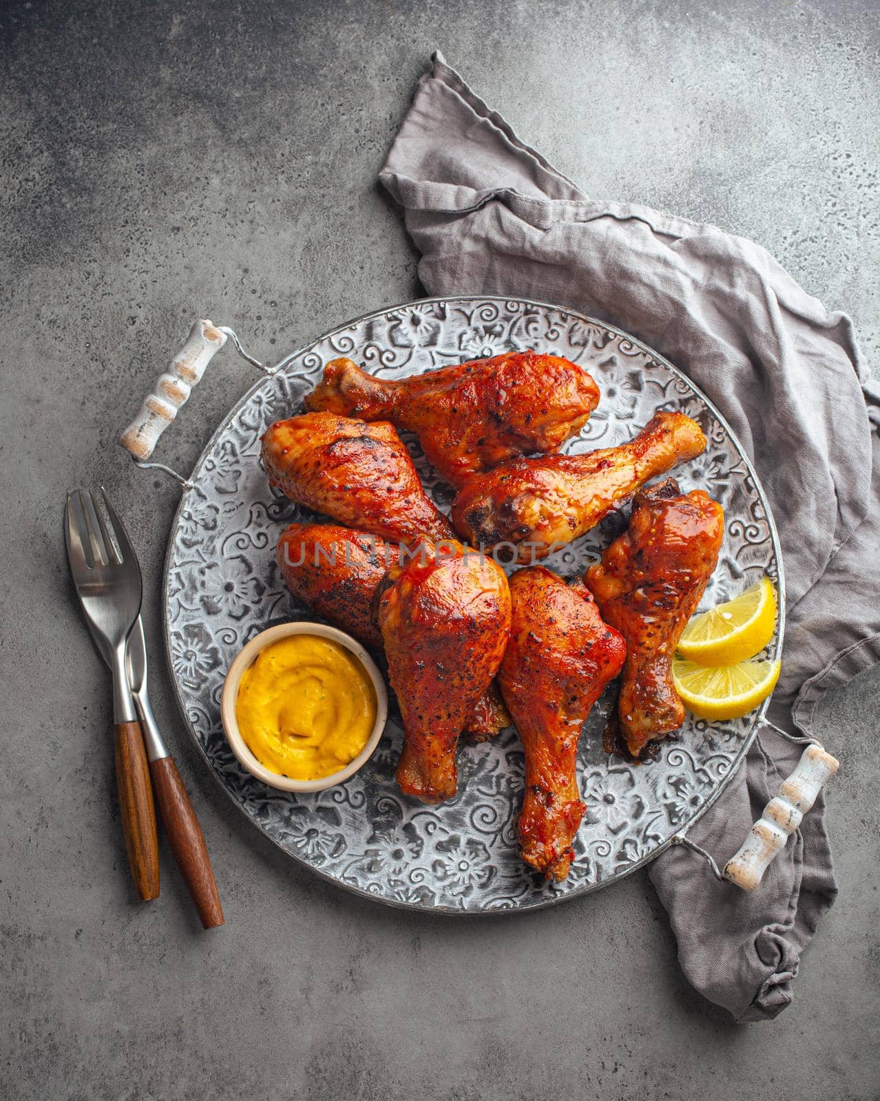 Traditional Indian dish Tandoori chicken legs served with exotic yellow sauce and lemon wedges on rustic aluminum plate on grey concrete background from above