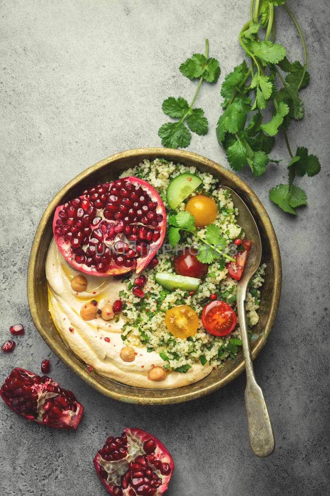 Rustic bowl with couscous salad with vegetables, hummus and fresh cut pomegranate. Middle eastern or Arab style meal with seasonings and fresh cilantro. Healthy Mediterranean dinner, toned image.