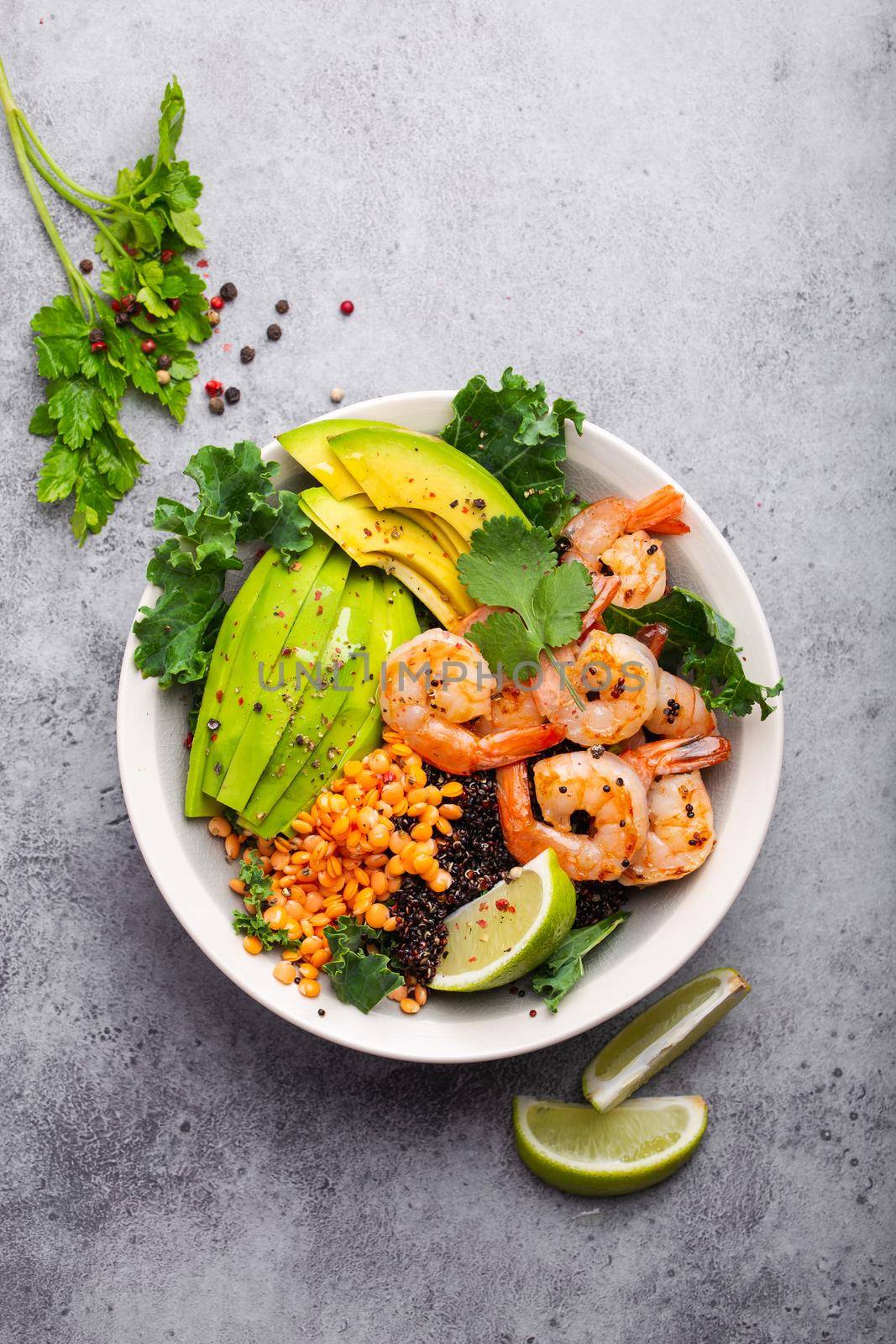 Close-up, top view of salad bowl with shrimps, avocado, fresh kale, quinoa, red lentils, lime and olive oil on gray stone background. Lunch bowl, healthy clean eating, dieting or nutrition concept