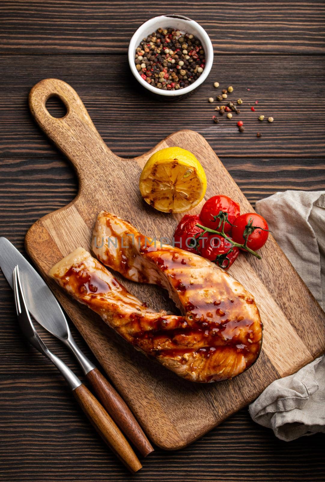 Cooked grilled salmon steak glazed with teriyaki sauce, cherry tomatoes and lemon served on cutting board on rustic dark wooden background from above