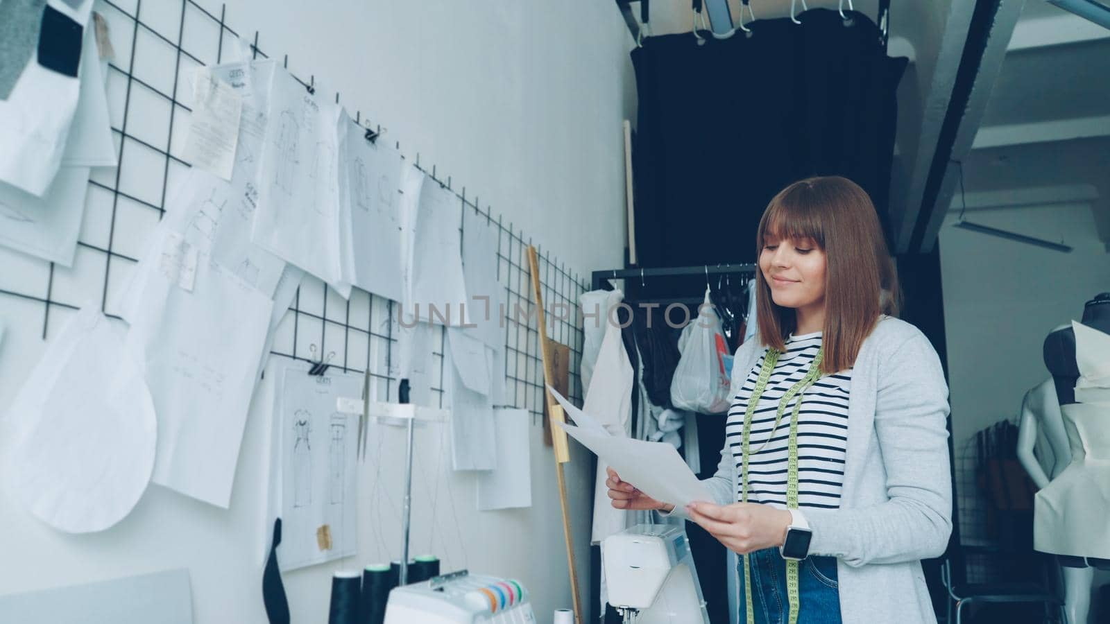 Young creative clothes designer is looking at sketches then hanging them on wall above studio table beside other drawings. Loft style studio and fashionable garments are visible.