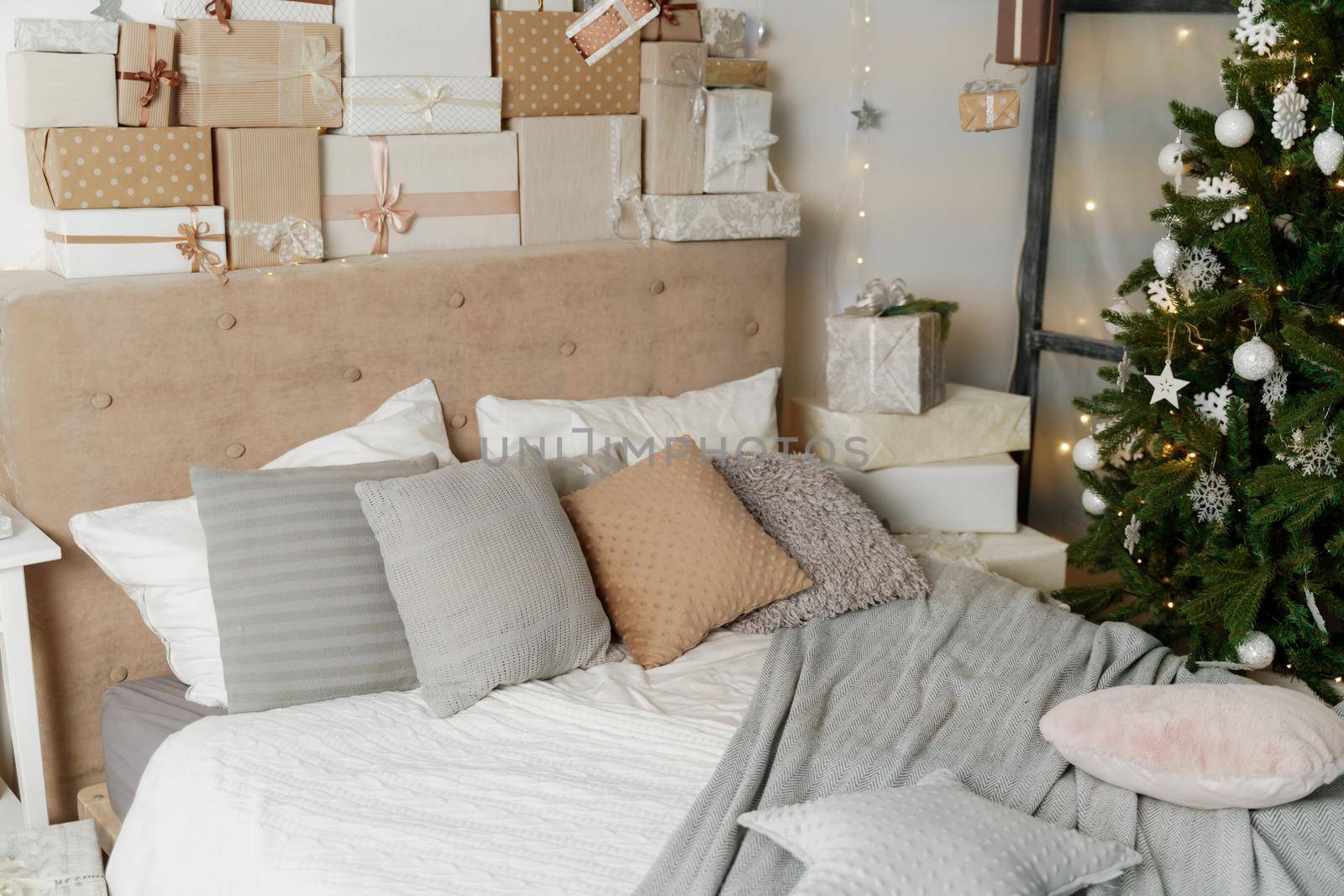 Pile of Christmas gifts in colorful wrapping with ribbons against a white wall with silver stars. Christmas tree in the room