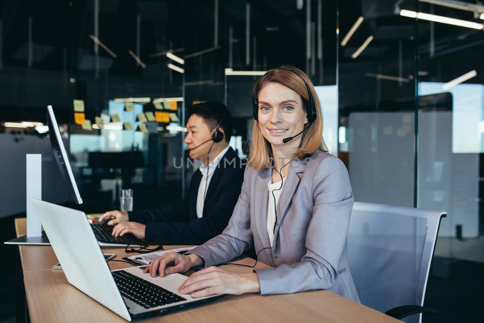 Portrait of a beautiful blonde woman, a call center employee looks at the camera and smiles, uses a headset for video communication, a team of online support operators