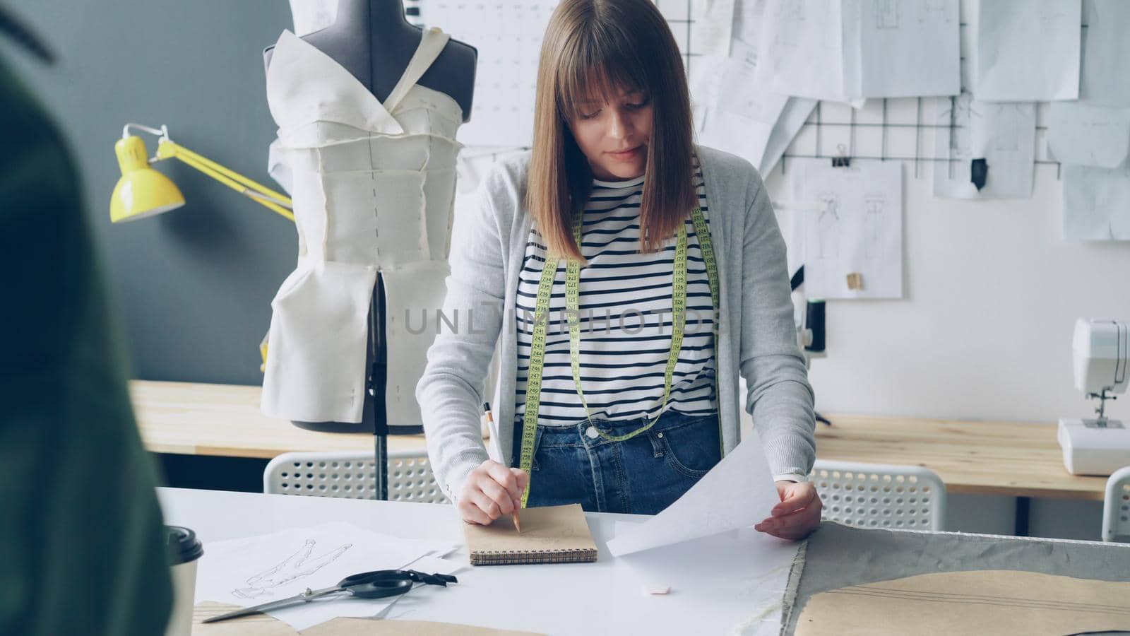 Professional dressmaker is making notes in notepad and looking at clothing sketch while working at studio table. Many drawings, dummy and sewing machine in background. by silverkblack