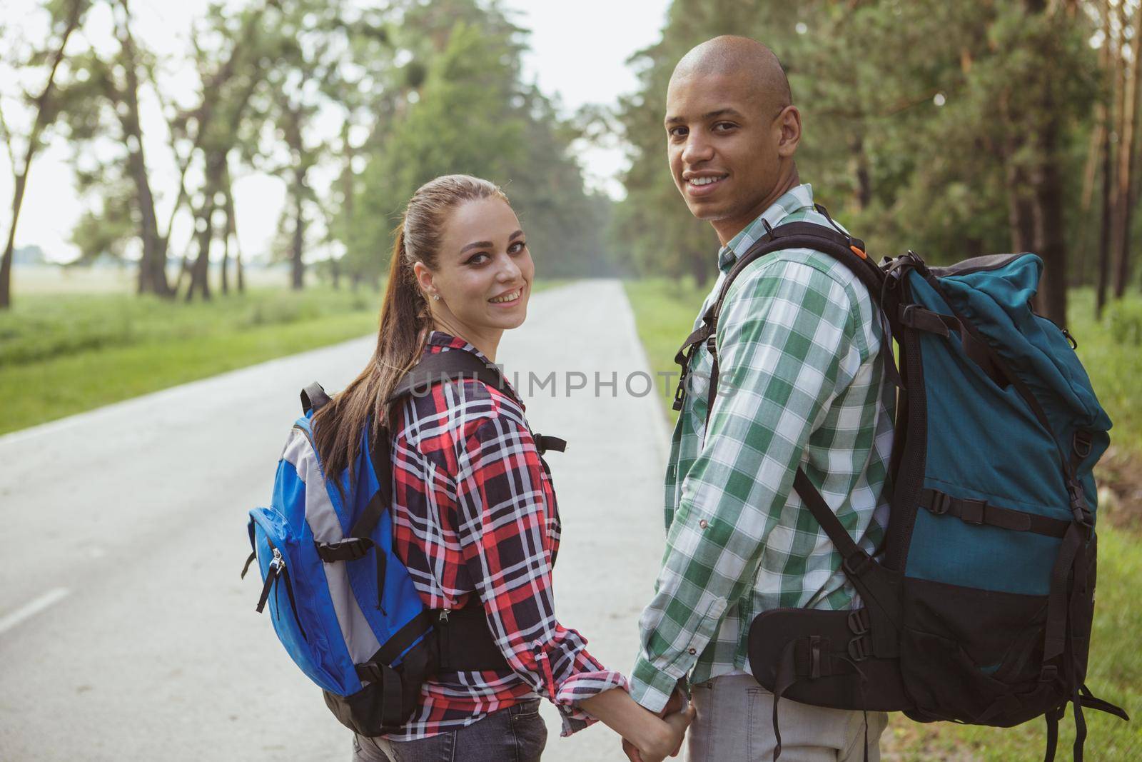 Happy young multiracial couple holding hands while backpacking together, smiling to the camera. Lovely mixed couple walking in countryside road near forest, travelling together