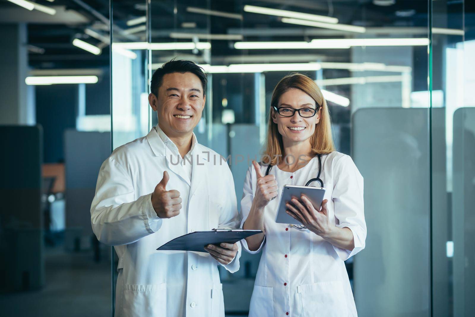 Team of two doctors asian man and woman looking at camera and smiling holding thumbs up working in modern clinic