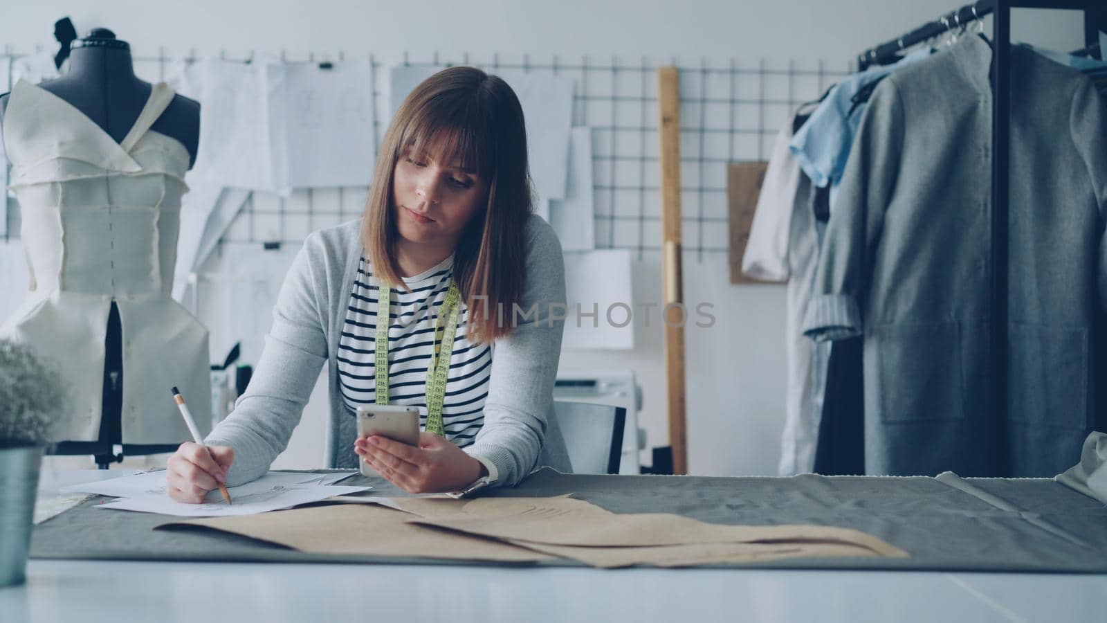 Creative clothing designer is looking at smartphone and drawing sketch while working in modern tailor shop at sewing table. Woman is busy and involved in process. by silverkblack
