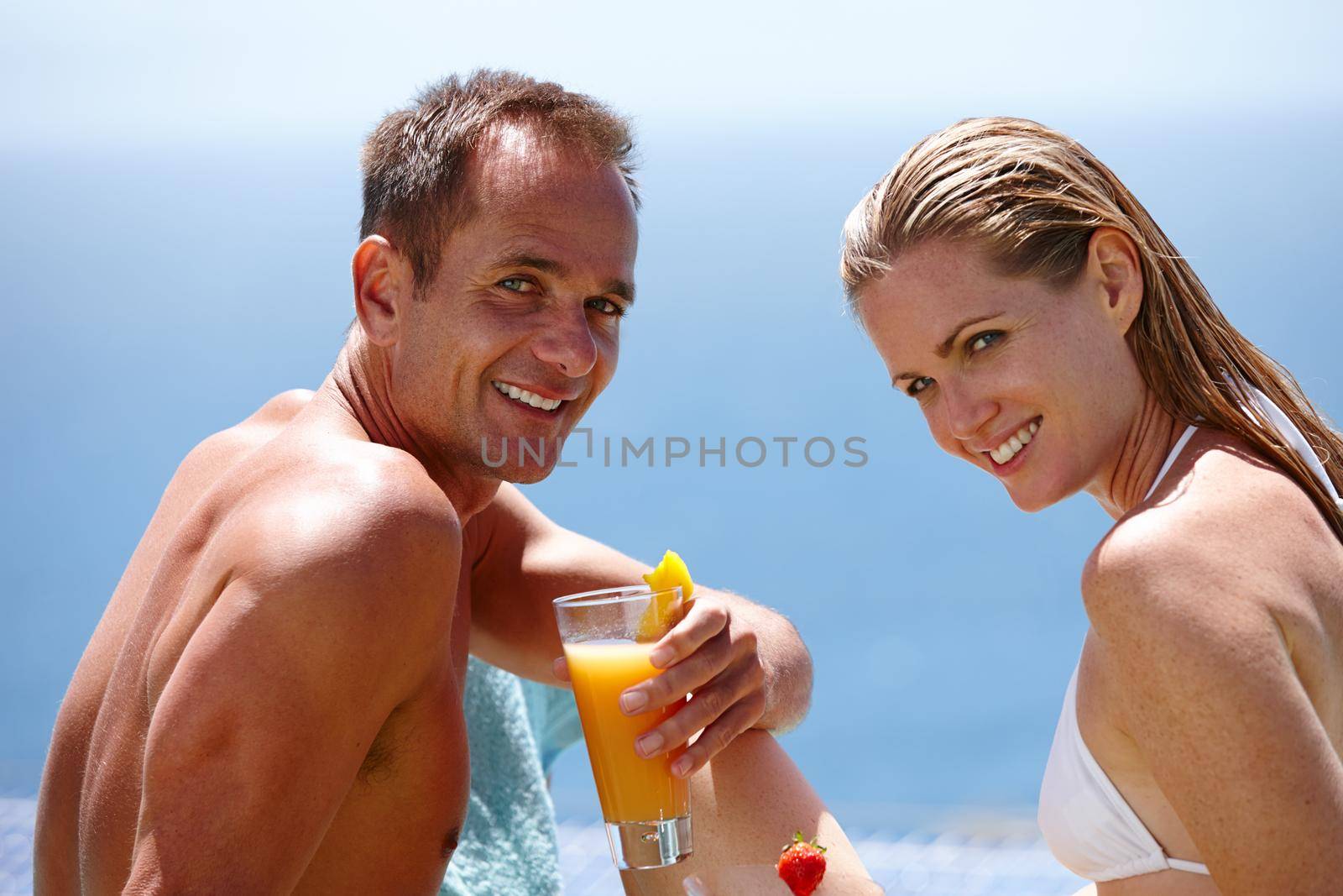 Romance and relaxation in the sunshine. Portrait of a smiling couple in their swimwear enjoying a drink outdoors. by YuriArcurs