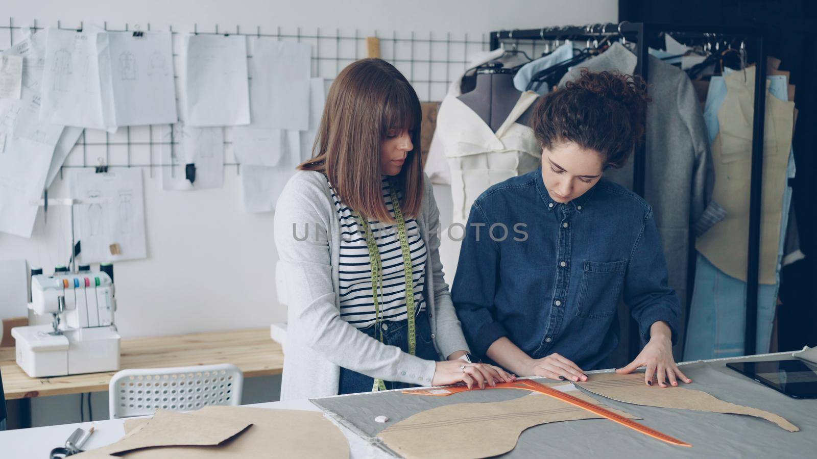 Experienced confident seamstress is teaching her assistant to outline clothing patterns on fabric. Young woman is focused on process, watching carefully and asking questions.