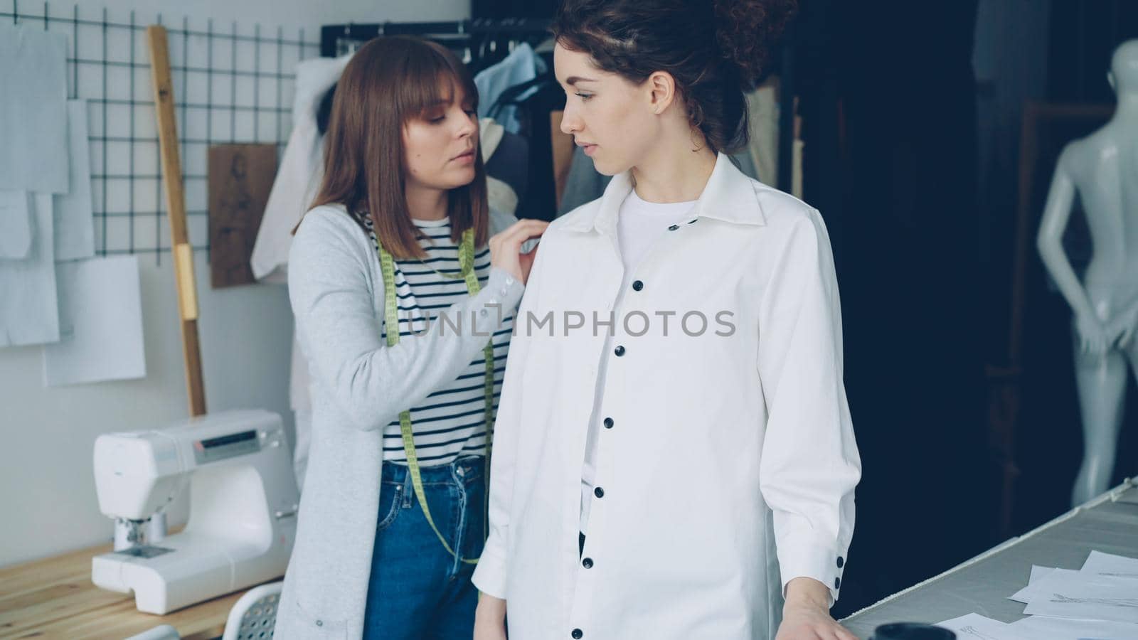 Pretty customer is trying on tailored shirt and sharing her opinion while clothing designer is measuring and checking garment and taking to client in light studio. by silverkblack