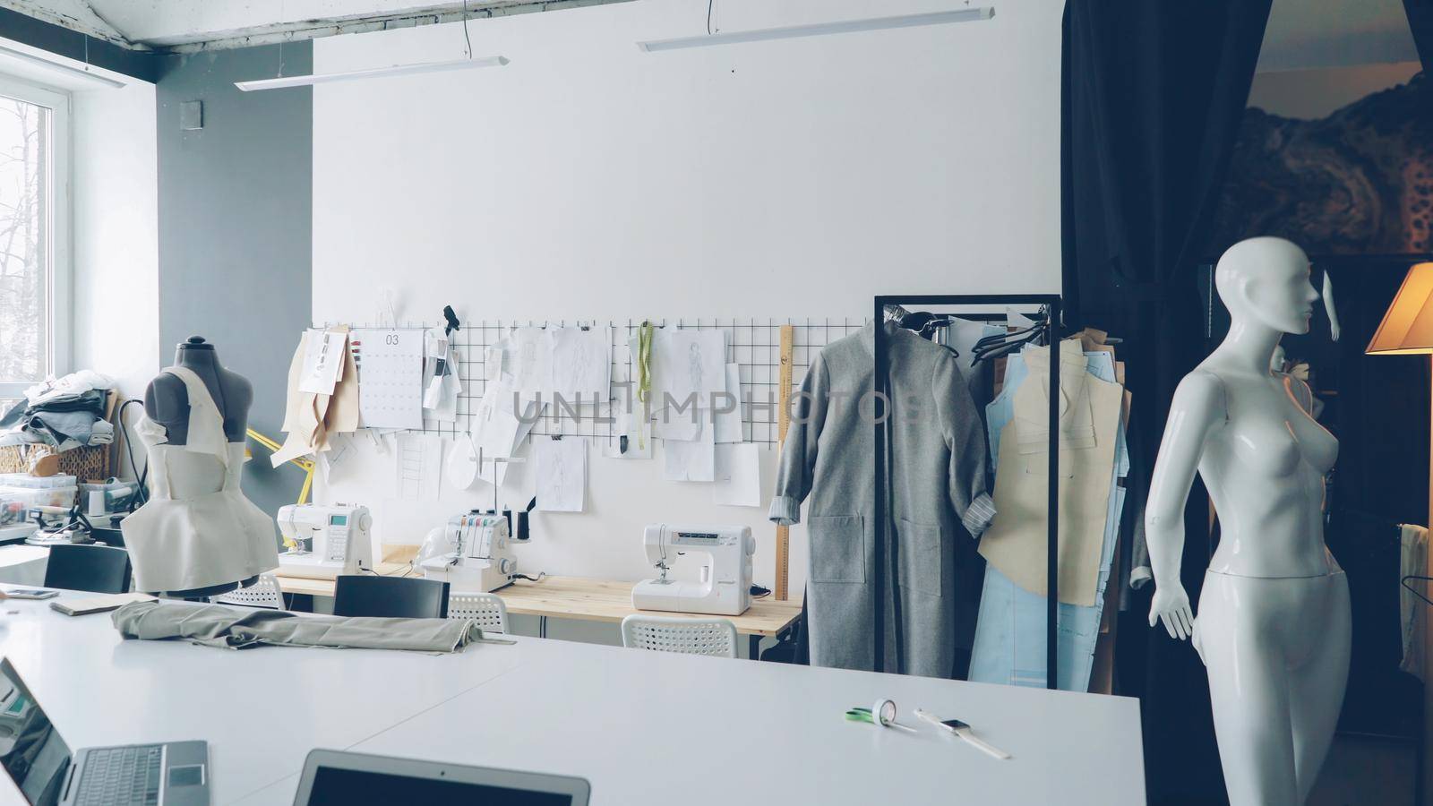 Light clothing design studio with large tailor's desk, mannequins, numerous sketches pinned on wall, sewing machines and half-finished garments on rails. by silverkblack