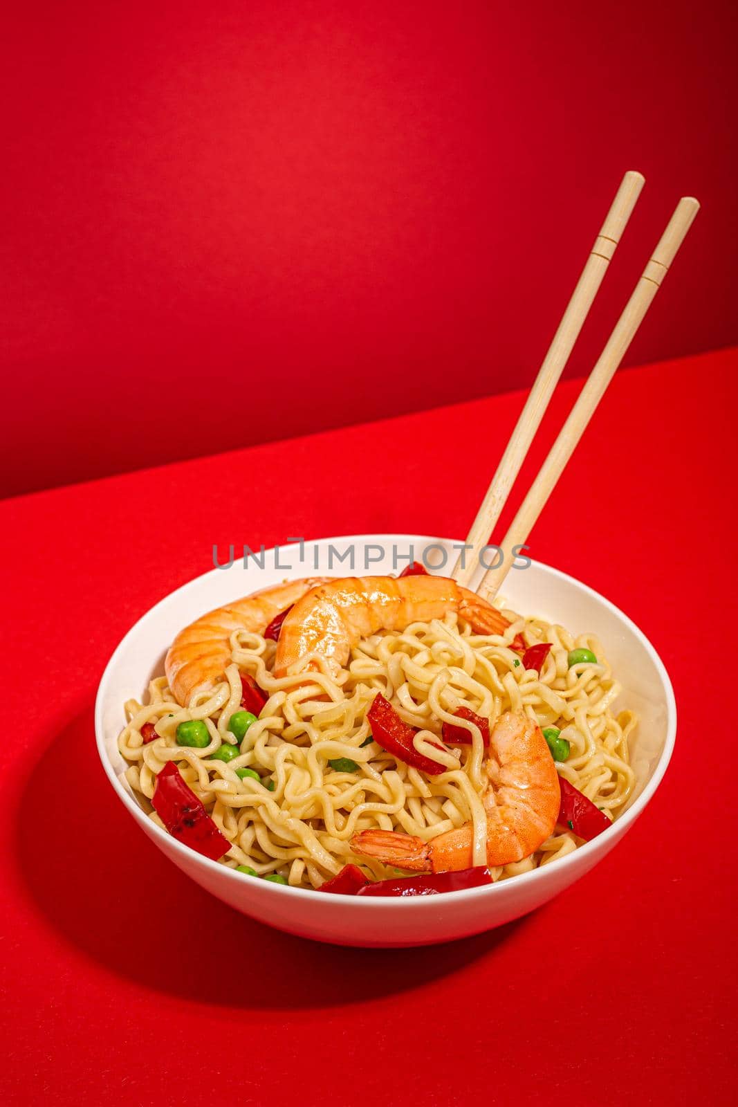 Asian Chinese noodles with shrimps and vegetables in white bowl with wooden sticks on red minimal paper background, hard sharp shadows, minimal foodphoto concept