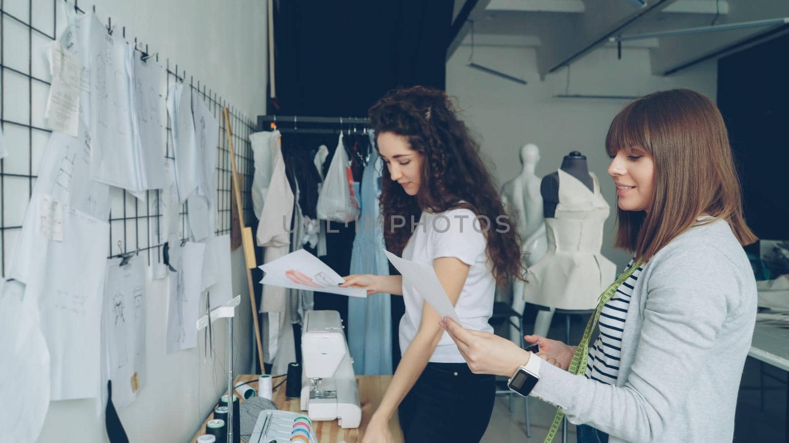 Attractive women clothing designers looking at garment sketches hanging on wall, discussing them, choosing new images for newest collection. Friendly informal atmosphere in studio.