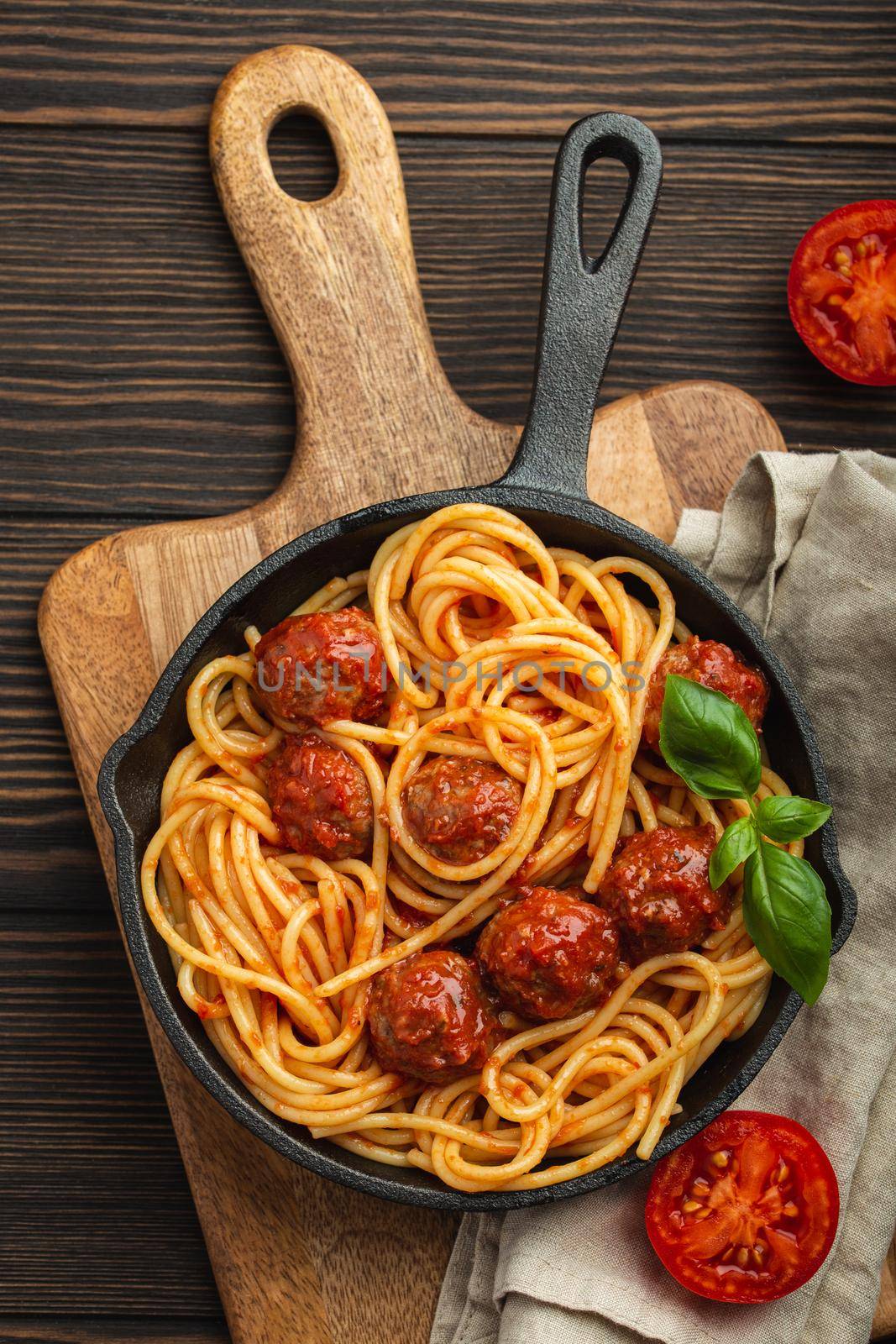 Top view of delicious pasta with meatballs, tomato sauce and fresh basil in cast iron rustic vintage pan served on cutting board, wooden background. Tasty homemade meatballs spaghetti