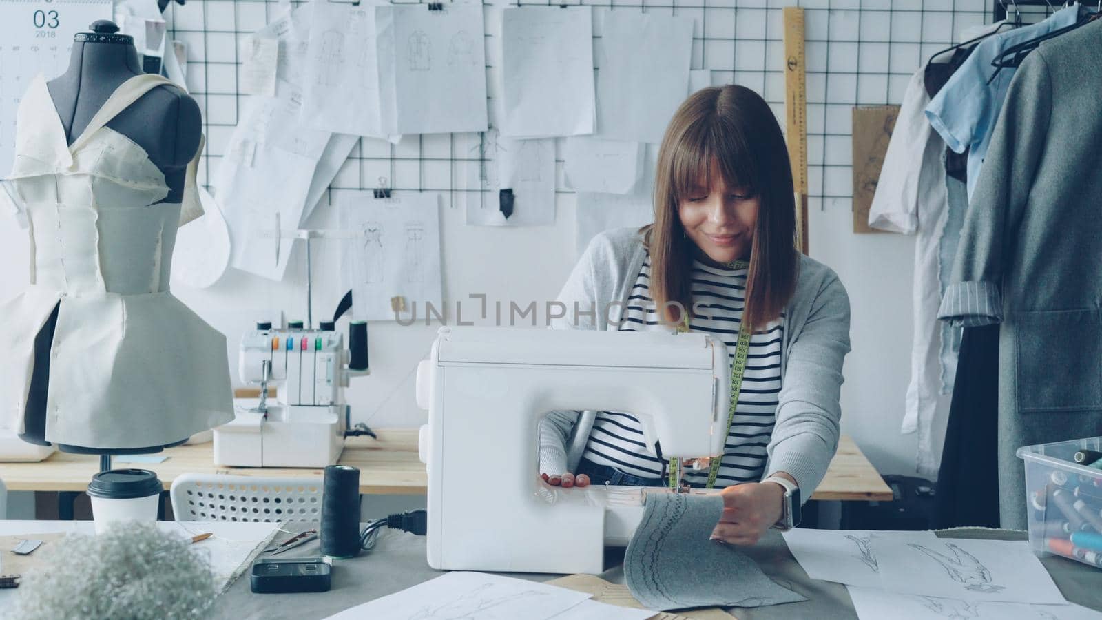 Young attractive dressmaker is working on sewing machine and looking at women's garment sketches in her studio at table. Modern equipment and fashionable garments in background. by silverkblack