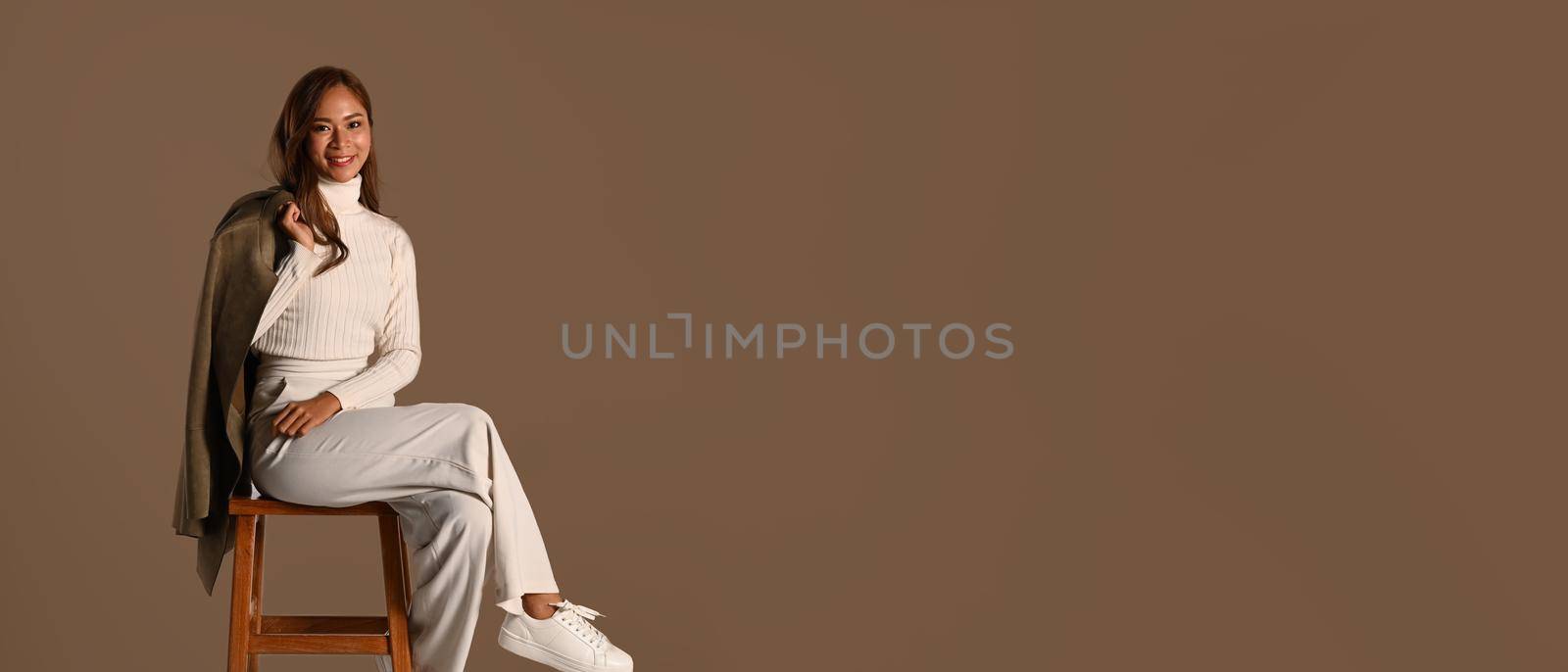 Panoramic shot of stylish woman holding trench coat sitting on wooden chair isolated on brown background. Autumn fashion concept by prathanchorruangsak