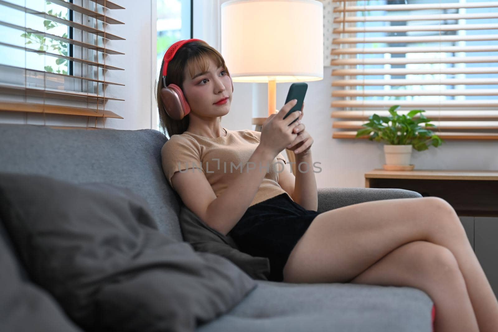 Beautiful young woman wearing headphone using mobile phone on couch at home by prathanchorruangsak