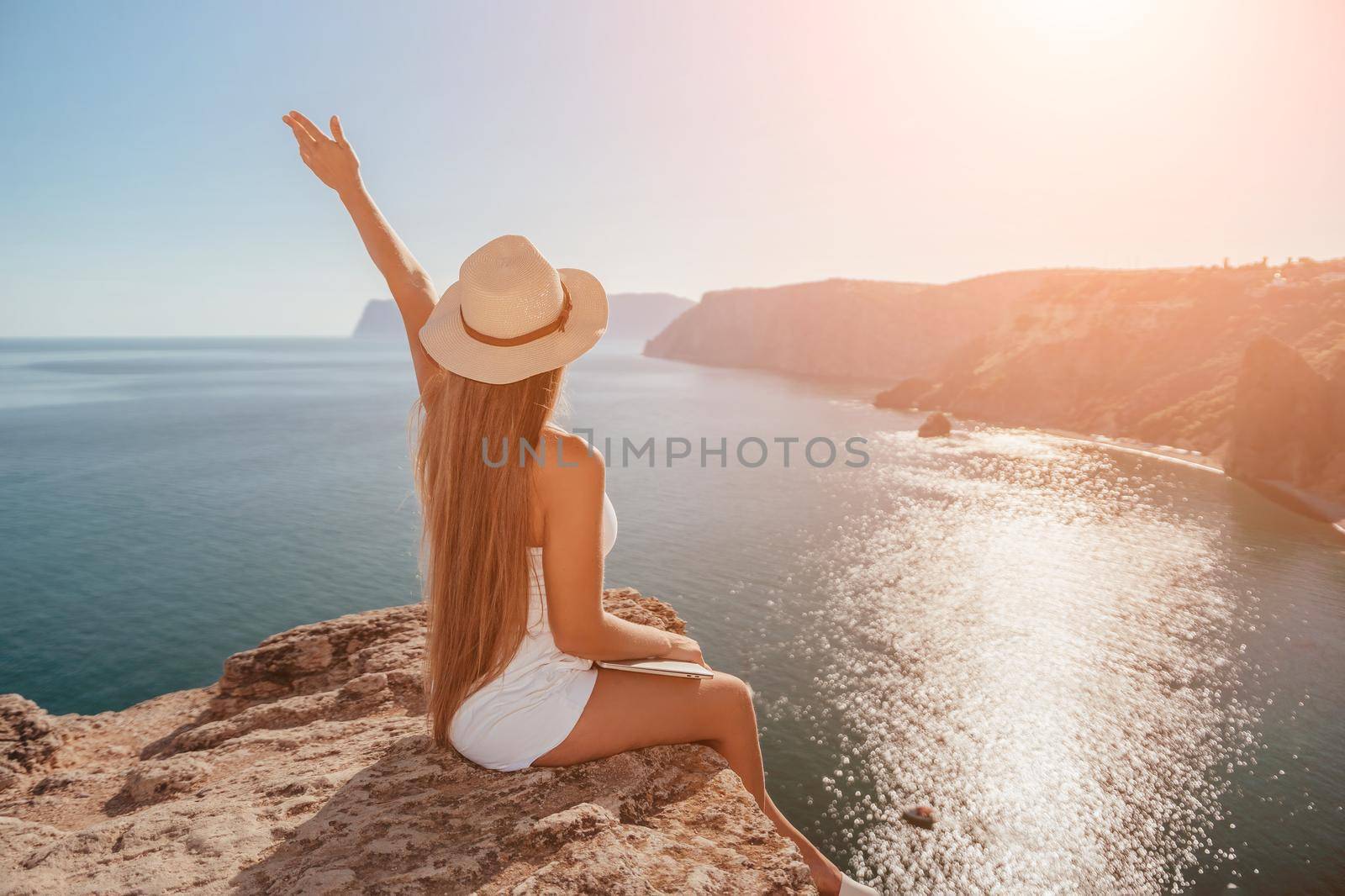 Digital nomad, woman in the hat, a business woman with a laptop sits on the rocks by the sea during sunset, makes a business transaction online from a distance. Freelance, remote work on vacation. by panophotograph