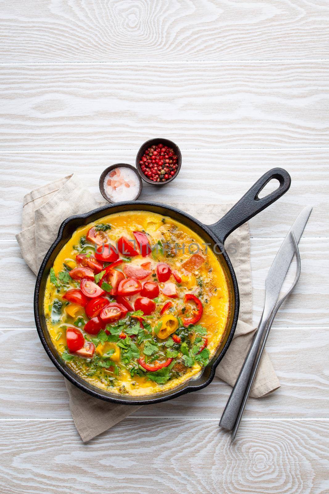 Healthy frittata in cast iron pan with fried beaten eggs and seasonal vegetables on white rustic wooden background. Italian omelette with organic spinach, bell pepper, tomatoes, from above