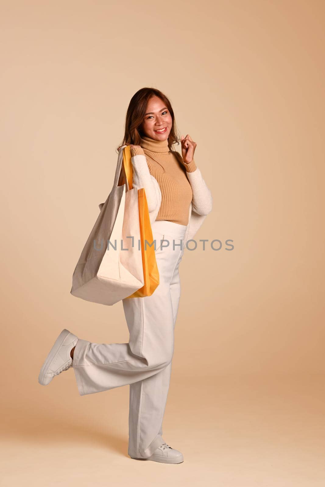 Full length of beautiful woman in warm sweater holding shopping bags standing on beige background. Black Friday concept by prathanchorruangsak