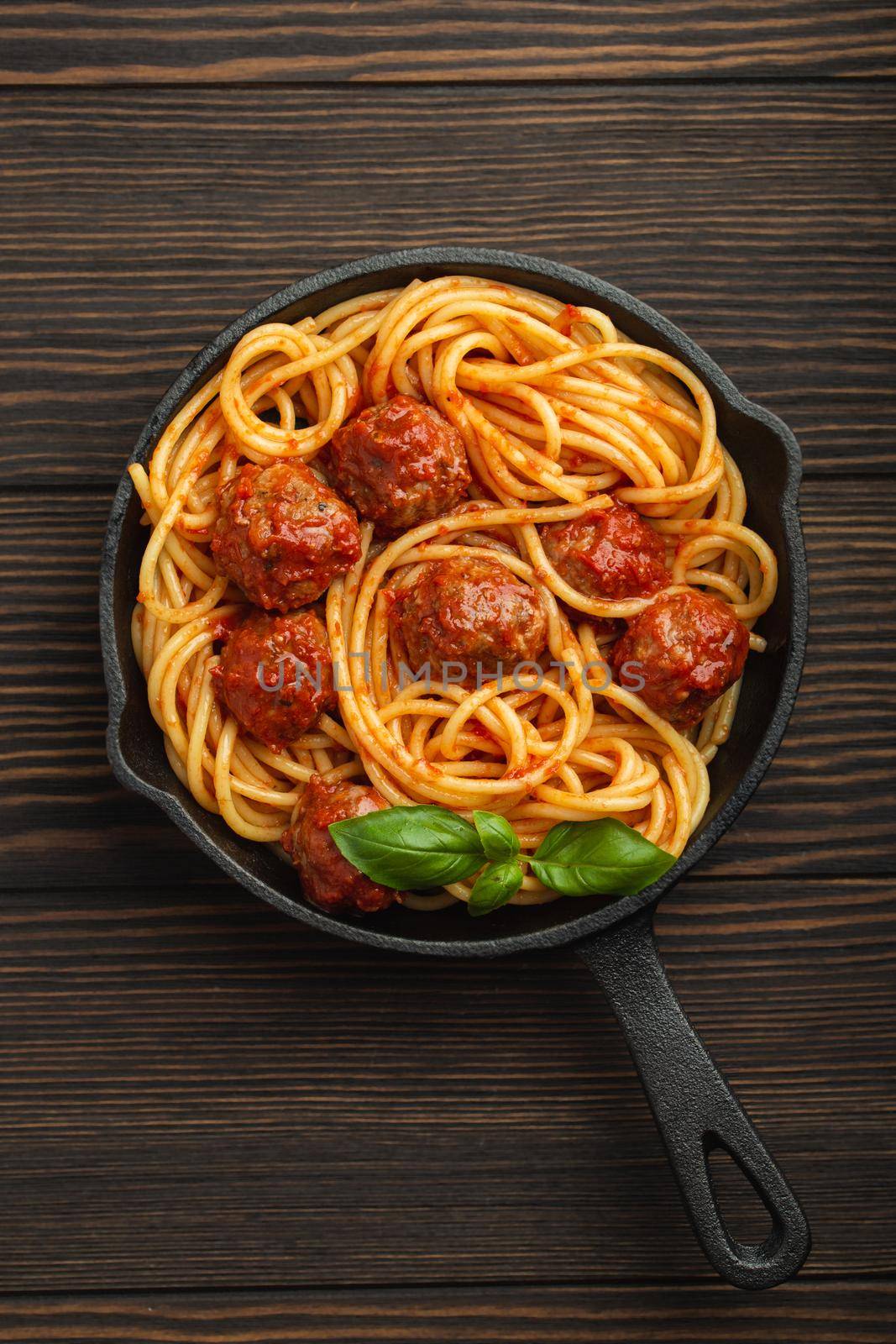 Top view of delicious pasta with meatballs, tomato sauce and fresh basil in cast iron rustic vintage pan served on cutting board, wooden background. Tasty homemade meatballs spaghetti