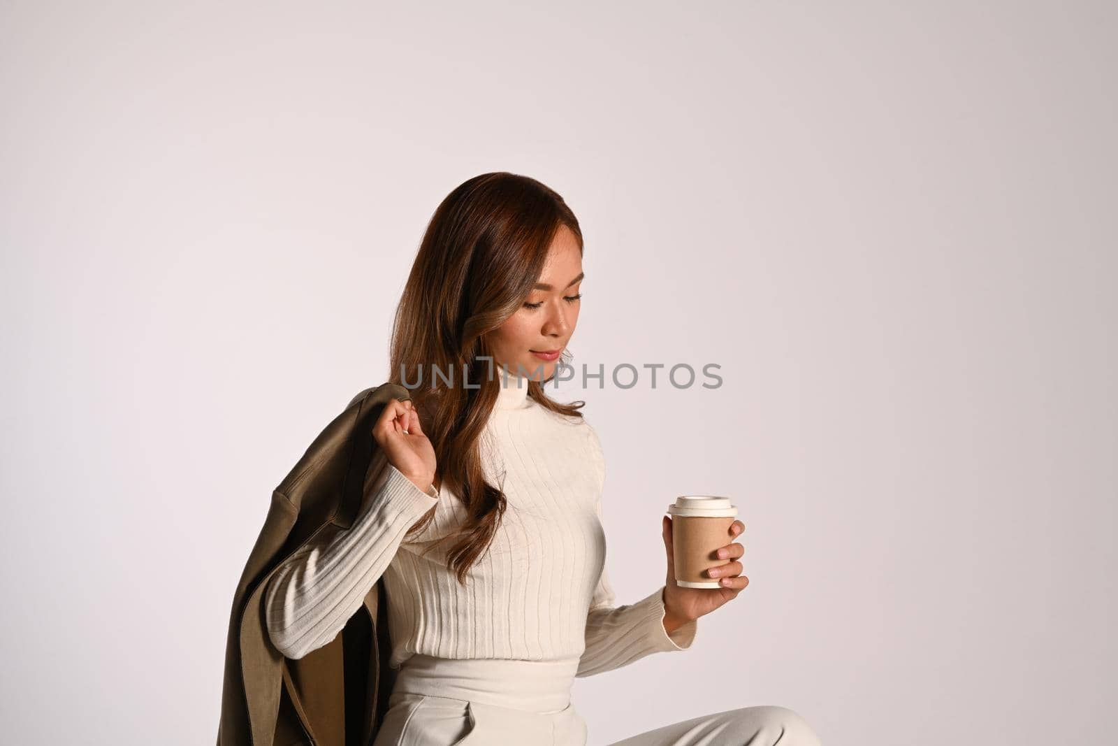 Young beautiful lady in fashionable autumn outfit holding paper cup sitting over light background. Autumn and Winter concept.