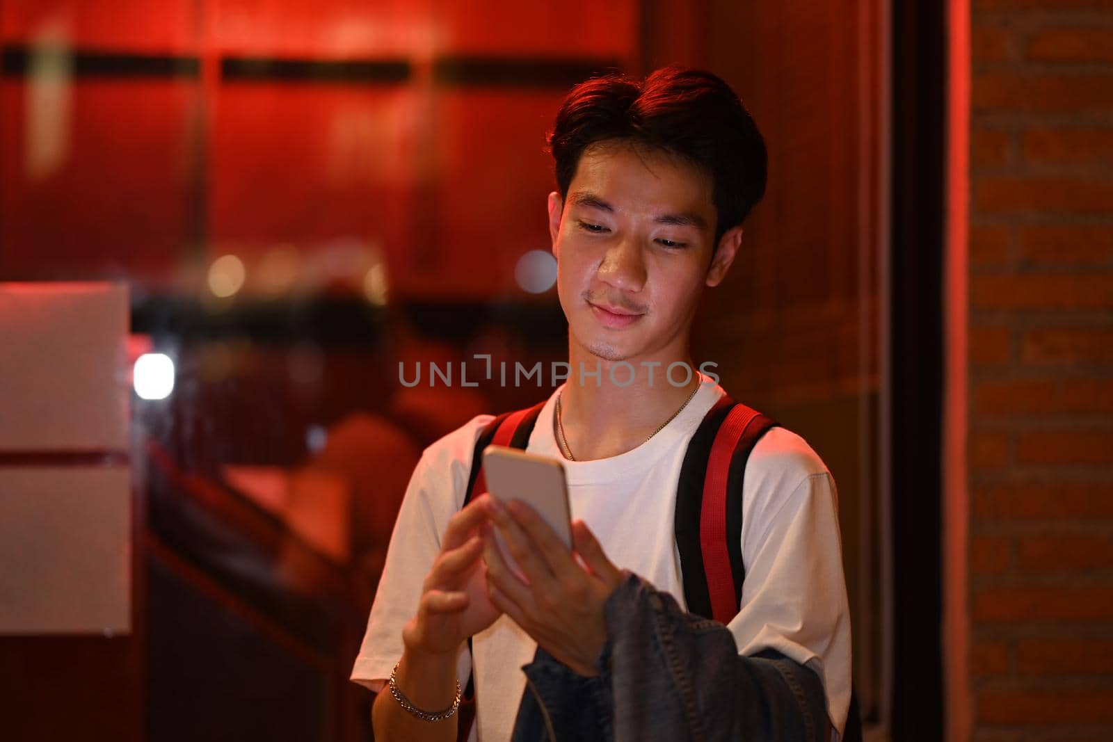 Asian male traveler standing on night city street near showcase and using mobile phone. Night life concept by prathanchorruangsak