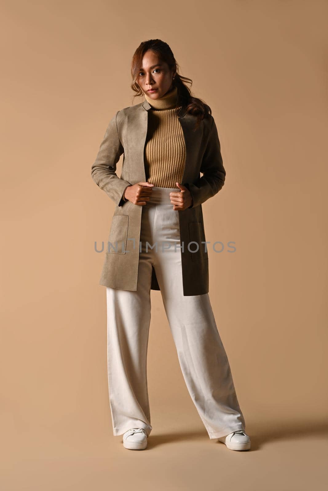 Full length portrait of fashionable woman in trench coat standing on beige background. Autumn fashion and beauty concept by prathanchorruangsak