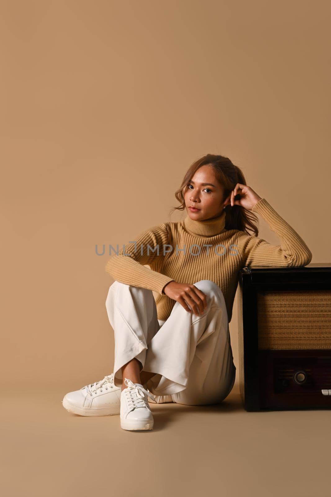 Portrait of fashionable woman wearing trendy sweater sitting near a vintage radio on beige background. Autumn fashion concept.