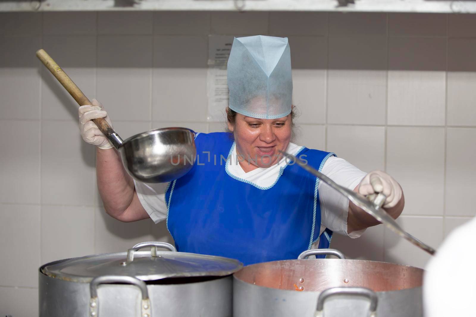 A woman cook in an industrial kitchen with a large ladle among metal pans. Chef at the hospital
