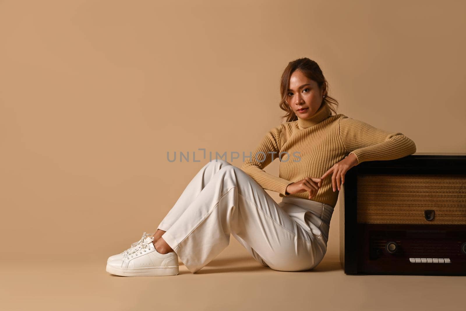 Fashion studio photo of young woman posing near a vintage radio over on beige background. Autumn fashion and beauty concept.