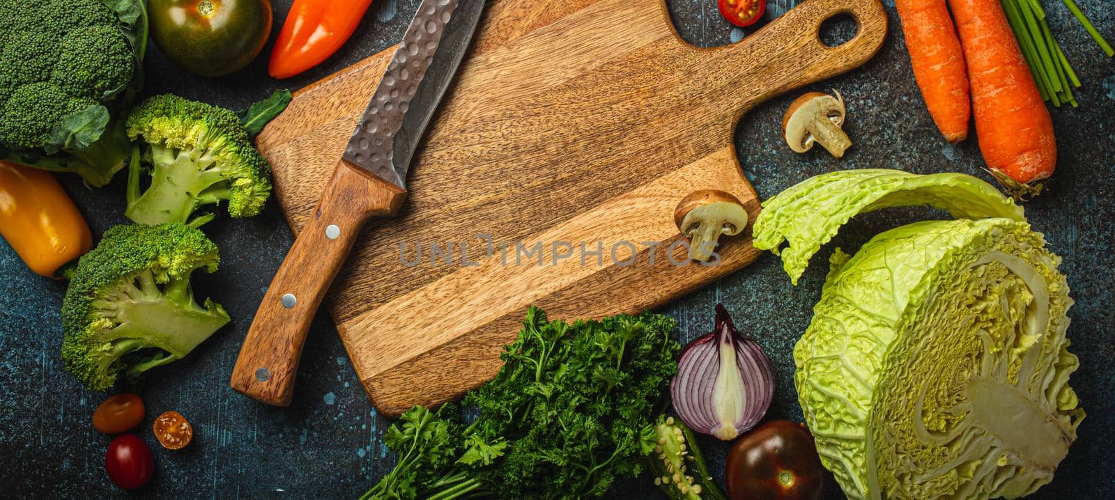 Assorted fresh vegetables on rustic concrete table with wooden cutting board in center and kitchen knife by its_al_dente