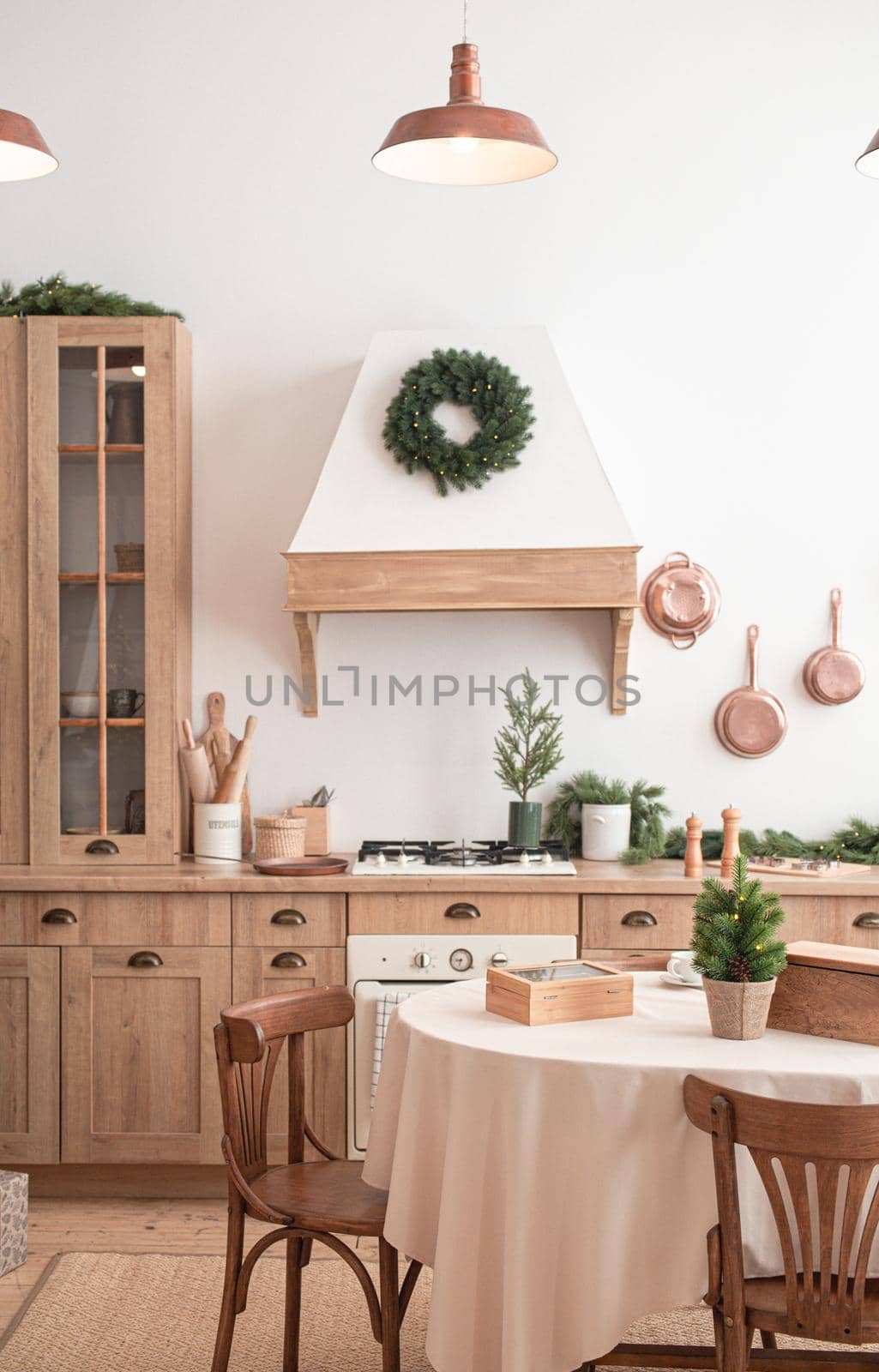 Modern, light, festive, cozy kitchen interior with Christmas and New Year decorations, kitchen table, utensils, copper pans on wall and big Christmas tree with presents, winter holidays concept