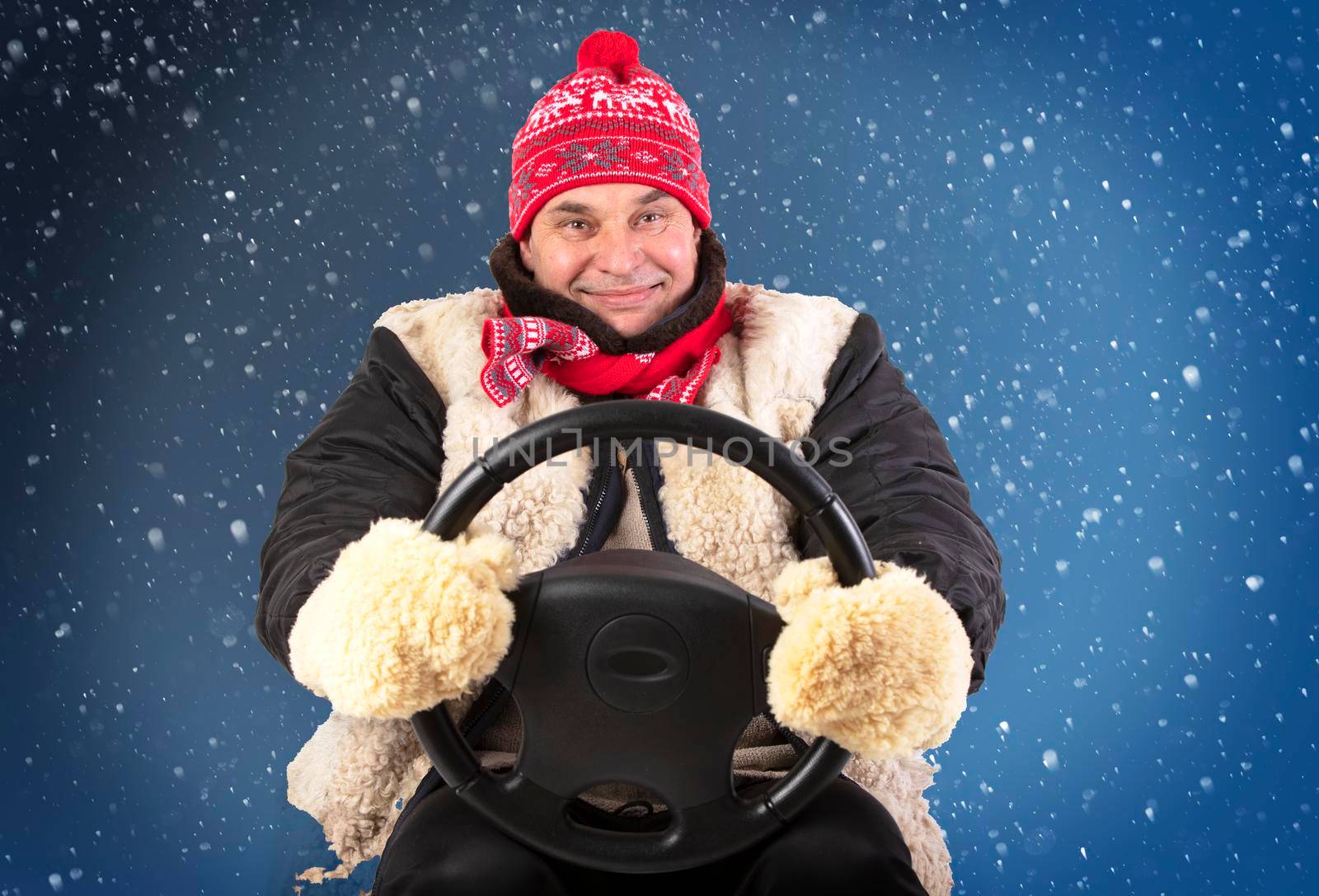 Funny adult man in a hat and fur mittens holds a car steering wheel on a blue snowy background.