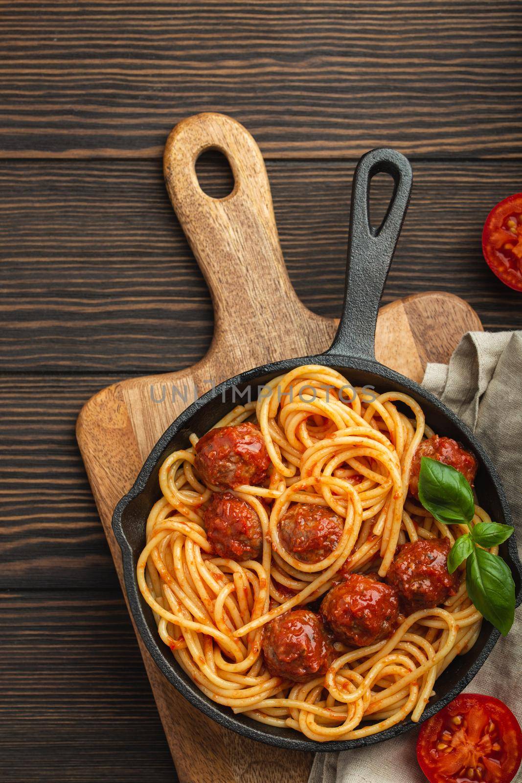 Top view of delicious pasta with meatballs, tomato sauce and fresh basil in cast iron rustic vintage pan served on cutting board, wooden background. Tasty homemade meatballs spaghetti