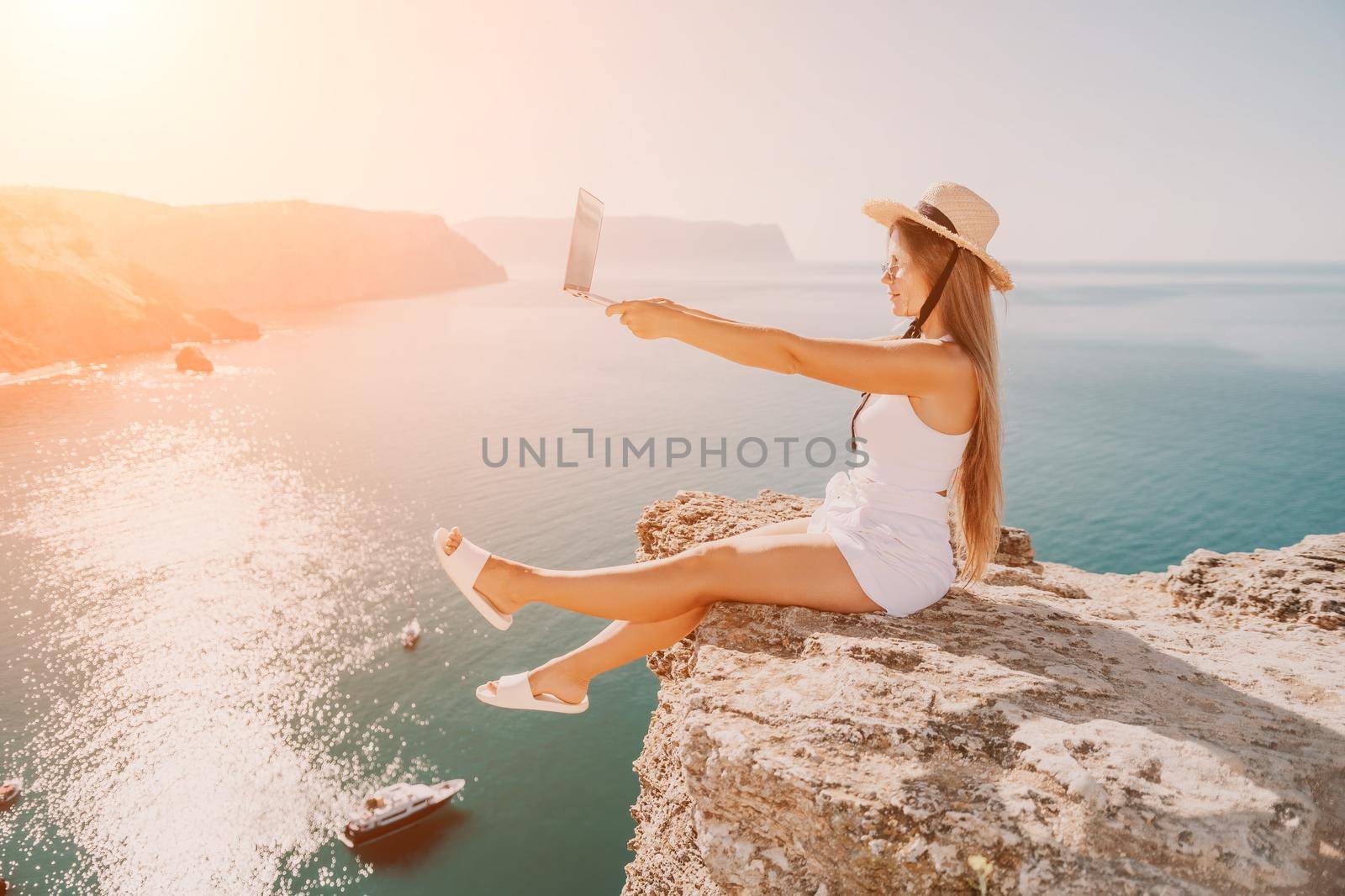 Digital nomad, woman in the hat, a business woman with a laptop sits on the rocks by the sea during sunset, makes a business transaction online from a distance. Freelance, remote work on vacation. by panophotograph