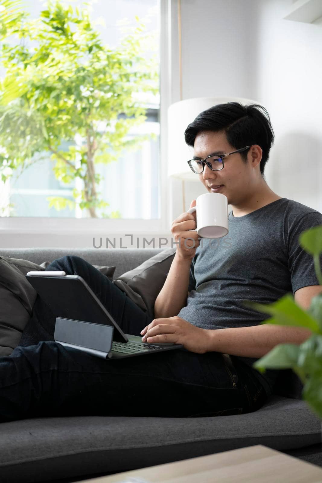 Young man holding coffee cup and working with computer tablet on sofa. by prathanchorruangsak