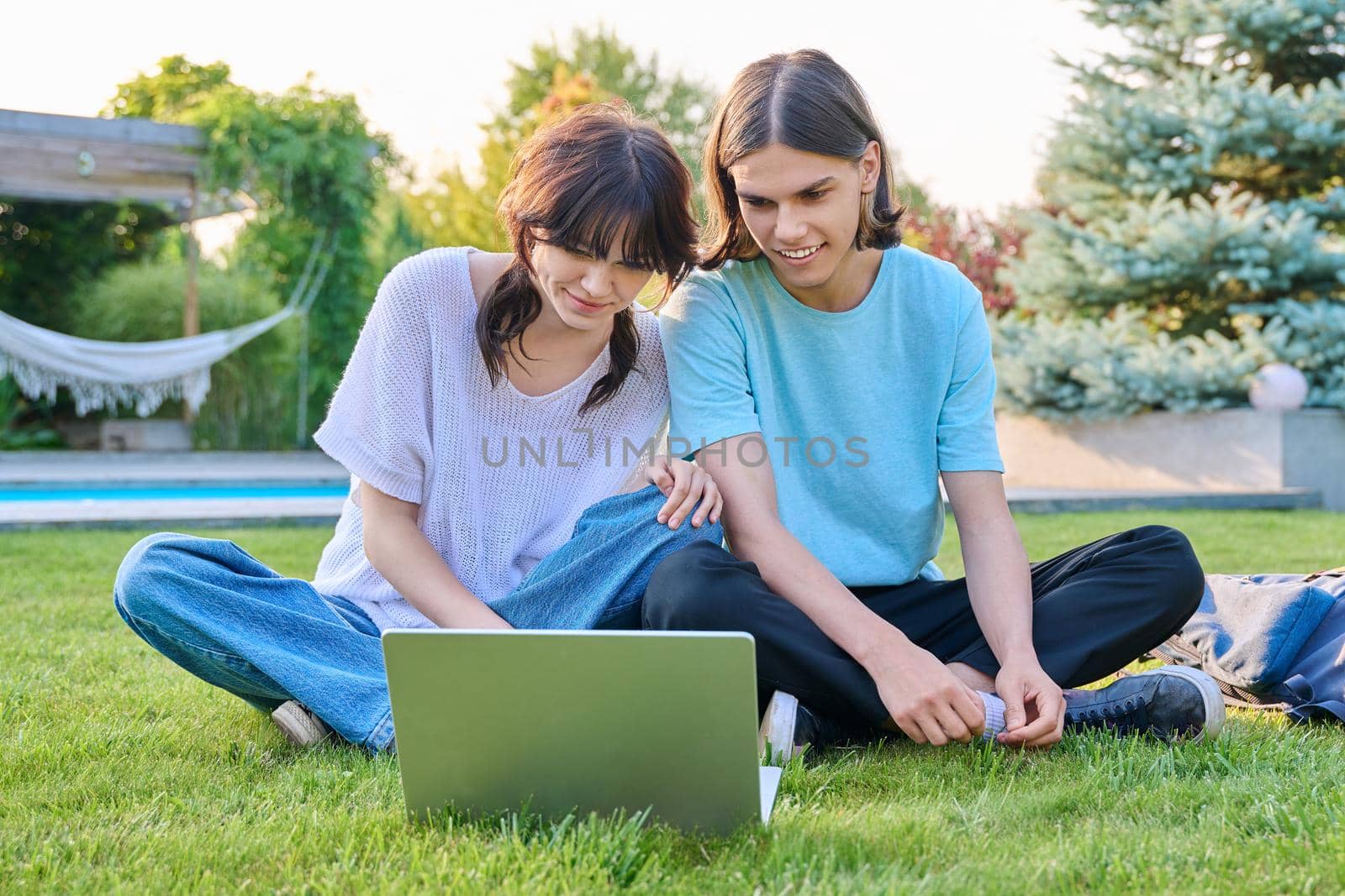 Two teenage friends of students sitting on grass with laptop, in backyard, guy and girl 18 years old study together. Friendship, youth, technology, high school, college, lifestyle concept
