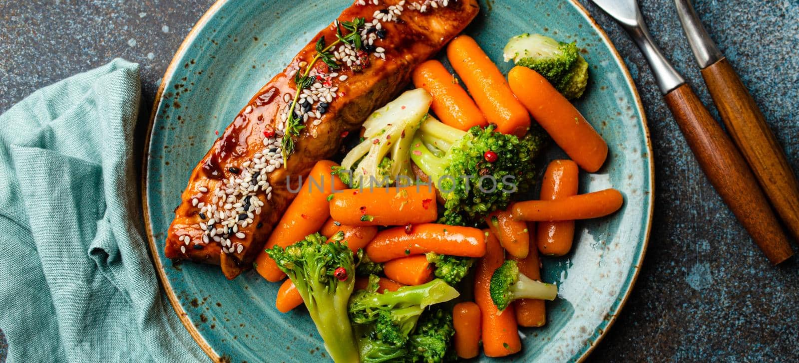 Grilled salmon fillet steak in teriyaki sauce with roasted vegetables carrot and broccoli on plate with fork and knife top view on rustic concrete background table, healthy diet and eating concept