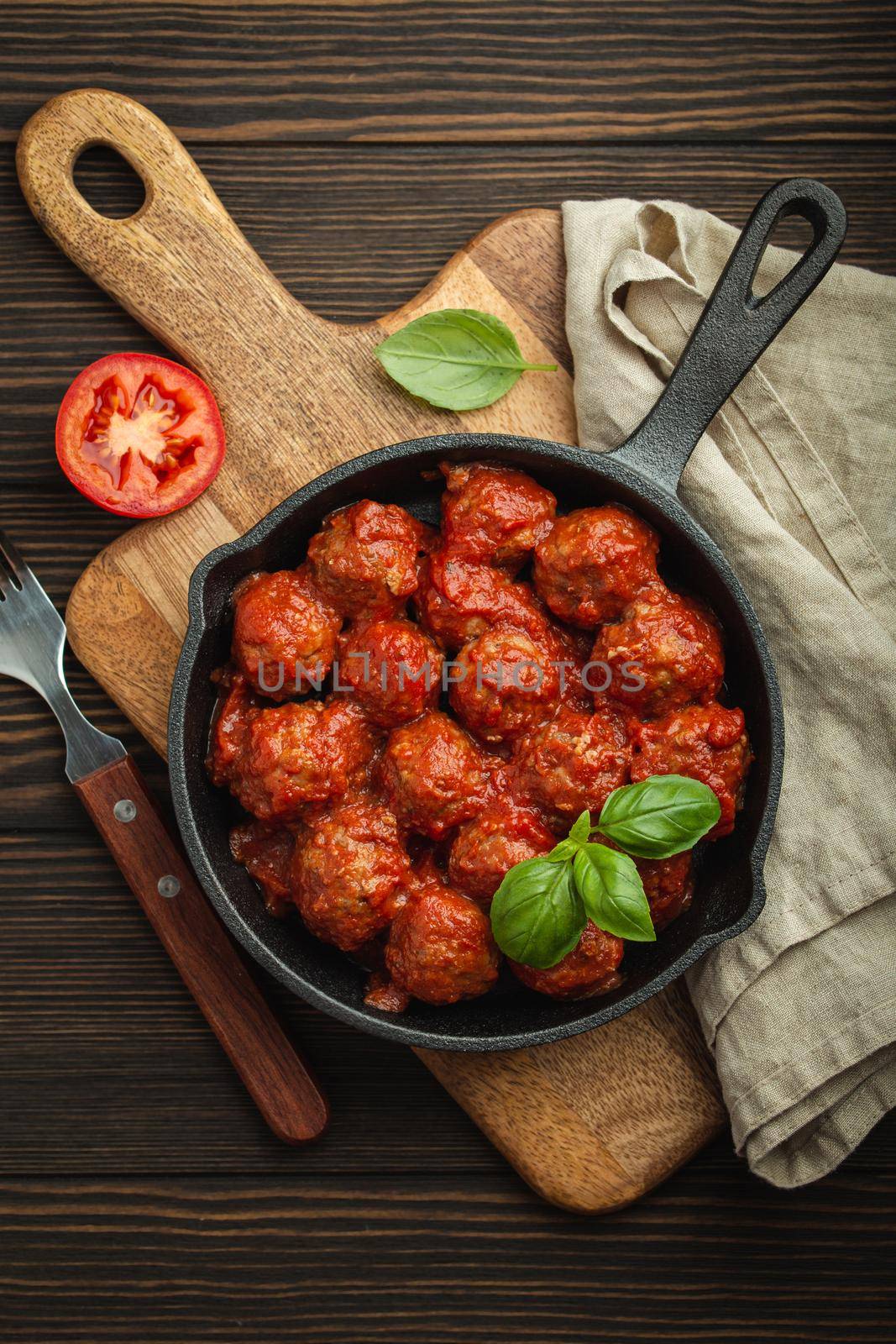 Top view of delicious meatballs with tomato sauce and fresh basil in cast iron rustic vintage pan served on cutting board, wooden background. Tasty homemade meatballs