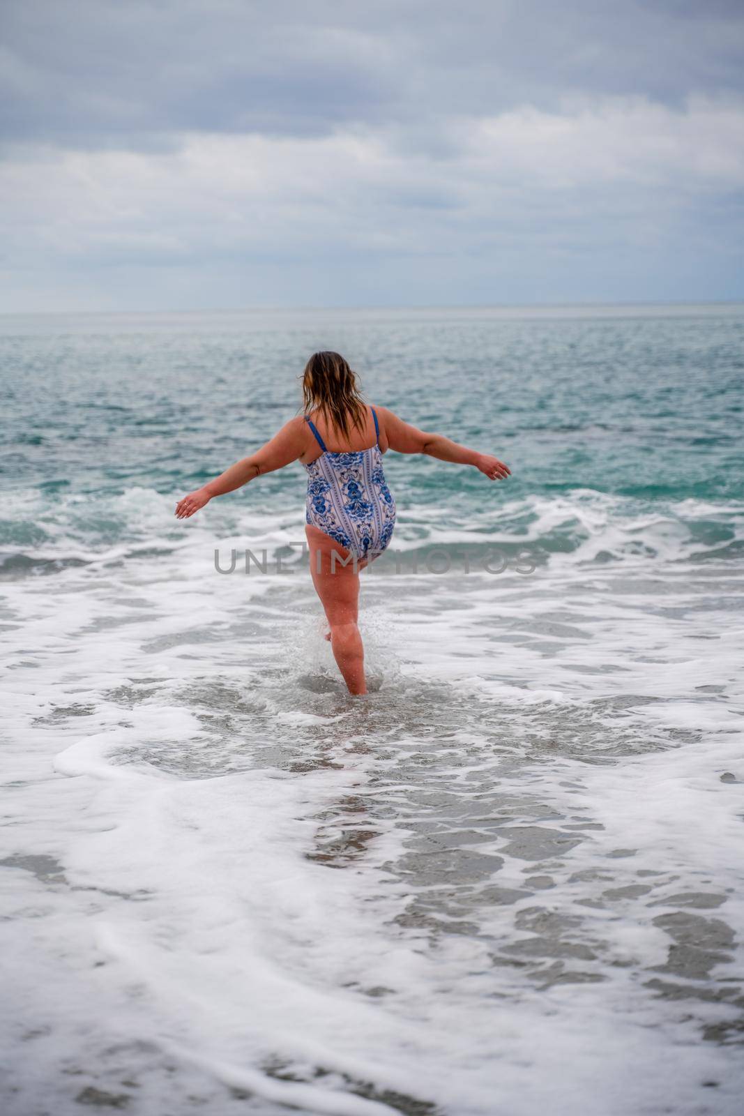 A plump woman in a bathing suit enters the water during the surf. Alone on the beach, Gray sky in the clouds, swimming in winter. by Matiunina