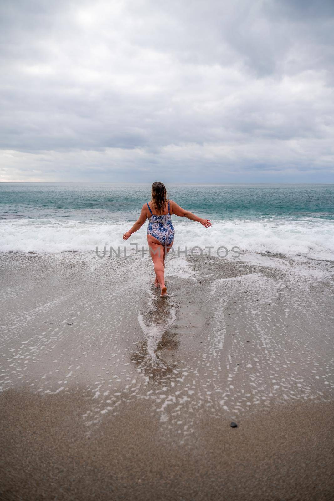 A plump woman in a bathing suit enters the water during the surf. Alone on the beach, Gray sky in the clouds, swimming in winter