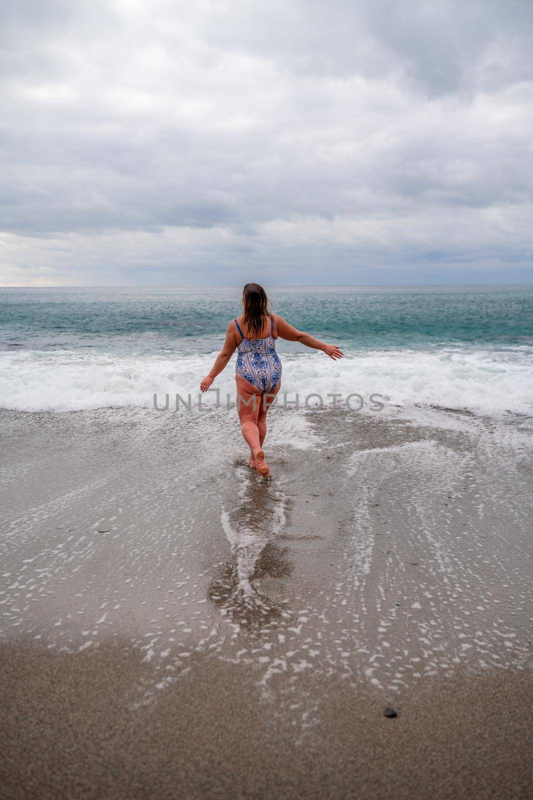 A plump woman in a bathing suit enters the water during the surf. Alone on the beach, Gray sky in the clouds, swimming in winter